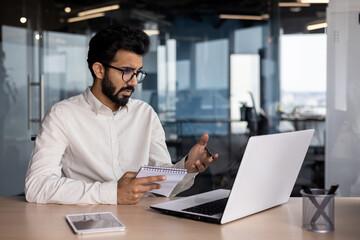 Fototapeta na wymiar Indian young male businessman working in office, sitting at desk holding notebook and talking on video call via laptop with clients and partners