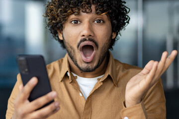Close-up portrait of a young Indian man looking shocked and surprised at the camera while holding a mobile phone and spreading his hands.
