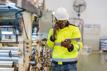 Half body portrait. African male engineer. Using a smartphone to talk to work, holding a laptop, wearing a vest and helmet, next to a working machine at a large company's plastic and steel industry.