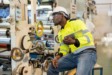 African male engineer Sit and check the system of the plastic and steel industry using a tablet and look at the machinery and gears. Wear a vest and helmet. To produce materials for the company