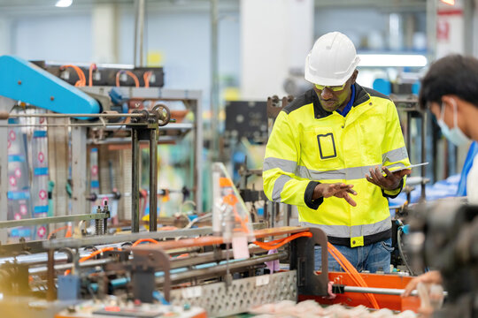African male engineer talking with employees while working, holding a laptop, wearing a vest, safety helmet. Around are working machines. Located in the plastic and steel industry.