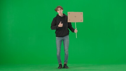 A male activist holds a blank poster and shows a thumbs up. A young man in casual clothes holds a placard on a green screen. Chromakey, advertisement, promo.