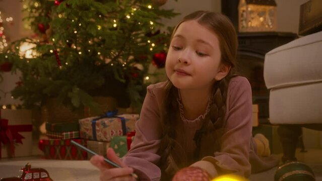 girl writes a letter with wish list to Santa Claus using color pencils laying on floor near Christmas tree.