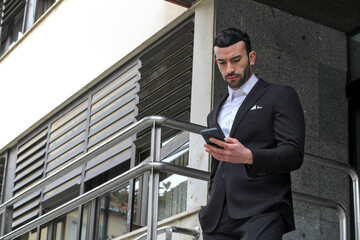 Young white man in suit standing stairs using the phone on the street, looking at the mobile, seen from below.