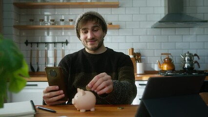 Young man with beanie sitting in modern kitchen at home while holding smartphone in his hand looking at the camera and puts couple of coin into the piggy bank - Powered by Adobe