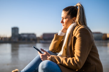 Sad woman holding phone while sitting by the river. 