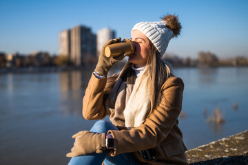 Beautiful woman in warm clothing enjoys  drinking coffee and resting by the river on a sunny winter...