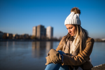 Sad woman in warm clothing sitting by the river on a sunny winter day.