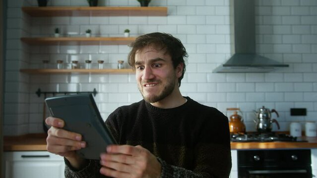 Young adult man standing in a modern kitchen, pressing the keys of a calculator frantically. He drops the calculator, goes mad and yelling screaming. Concept of anger during tax prep season