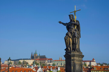 Statue of St. John the Baptist on Charles Bridge, with Hradcany castle and St. Vitus Cathedral in the background, in Prague, Czech Republic, in sunny day
