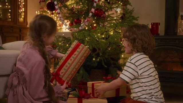 boy and girl run up to decorated christmas tree, take presents and shake them to guess what is inside, laugh smile and are excited of foretaste