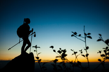 Silhouette of a hiker girl on a rock pedestal with hands up. Beautiful orange sunset on blue sky....