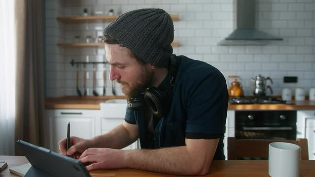 Focused Businessman With Beanie Working At Home In The Kitchen Working With His Tablet Computer, Taking Notes, Doing Research. Busy Freelancer Working On Modern Tech Notebook Device