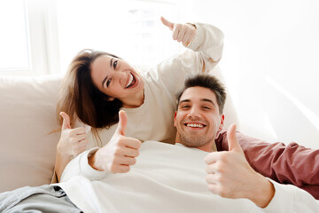 Portrait of attractive, handsome, smiling couple lying on comfortable sofa showing thumbs up