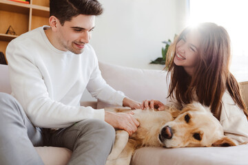 Beautiful couple, boyfriend and girlfriend petting dog, golden retriever lying on comfortable sofa