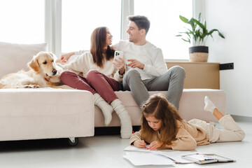 Smiling mother and father, woman holding mobile phone, dog lying on sofa daughter sitting on floor - obrazy, fototapety, plakaty