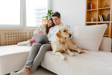 Happy young couple using laptop, man holding mobile phone, hugging dog. Online shopping concept - obrazy, fototapety, plakaty