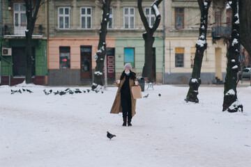 A young girl in a protective medical blue mask, a brown down jacket and a hat walks through a...