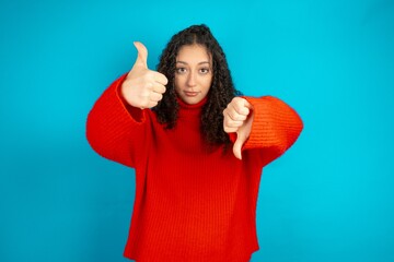 Beautiful teen girl wearing knitted red sweater over blue background showing thumbs up and thumbs down, difficult choose concept - obrazy, fototapety, plakaty