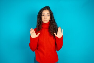 Serious beautiful teen girl wearing red knitted sweater pulls palms towards camera, makes stop...