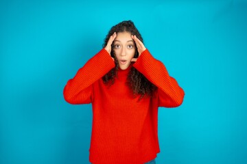 Beautiful teen girl wearing knitted red sweater over blue background with scared expression, keeps...