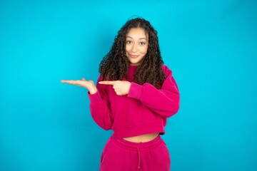 Beautiful young girl wearing pink tracksuit on blue background pointing and holding hand showing...