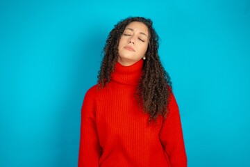 Beautiful teen girl wearing knitted red sweater over blue background looking sleepy and tired,...