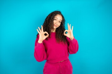 Beautiful young girl wearing pink tracksuit on blue background showing both hands with fingers in...