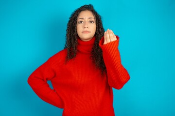 Beautiful teen girl wearing knitted red sweater over blue background angry gesturing typical...