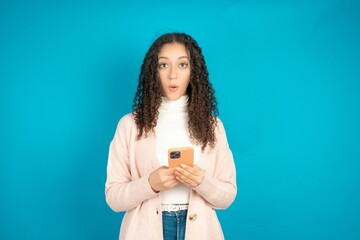 Shocked beautiful teen girl wearing brown cardigan opens mouth hold phone reading advert...