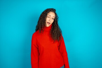 Beautiful teen girl wearing knitted red sweater over blue background winking looking at the camera...