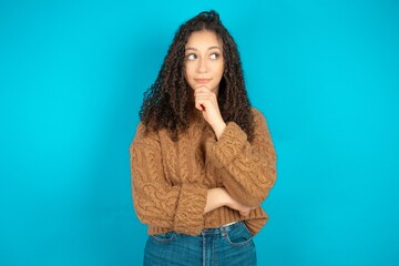 Thoughtful beautiful teen girl wearing brown knitted sweater holds chin and looks away pensively...