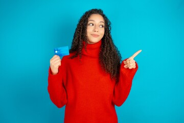 Curious smiling beautiful teen girl wearing red knitted sweater showing plastic bank showing finger copyspace