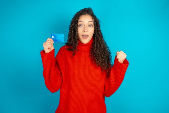 Photo of lucky impressed beautiful teen girl wearing red knitted sweater arm fist holding credit card. Celebrated