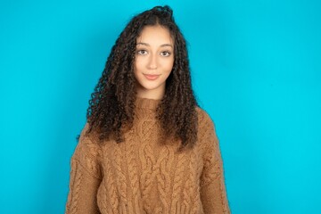 Beautiful teen girl wearing knitted sweater over blue background with nice beaming smile pleased...