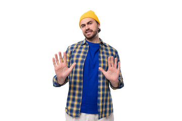 young doubtful european brunette man with a beard and mustache dressed in a yellow cap and a blue shirt shrugs his shoulders on a white background with copy space