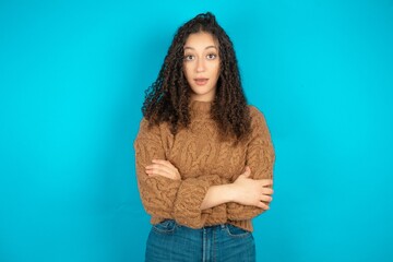 Shocked embarrassed Beautiful teen girl wearing knitted sweater over blue background keeps mouth...