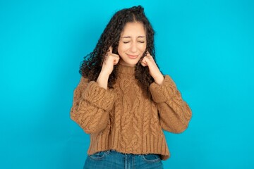 Happy Beautiful teen girl wearing knitted sweater over blue background ignores loud music and plugs...