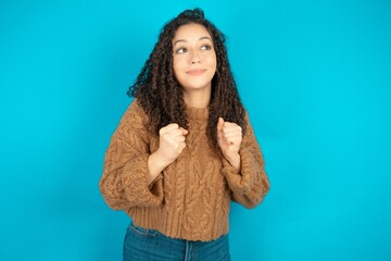 Beautiful teen girl wearing knitted sweater over blue background clenches fists and awaits for...