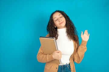 Overjoyed successful beautiful arab student carries notebooks raises palm and closes eyes in joy being entertained by friends
