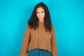 Portrait of dissatisfied Beautiful teen girl wearing knitted sweater over blue background smirks...
