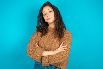 Beautiful teen girl wearing knitted sweater over blue background arms crossed look empty space billboard