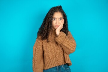 Sad lonely Beautiful teen girl wearing knitted sweater over blue background touches cheek with hand bites lower lip and gazes with displeasure. Bad emotions - Powered by Adobe