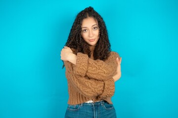 Charming pleased Beautiful teen girl wearing knitted sweater over blue background  embraces own...