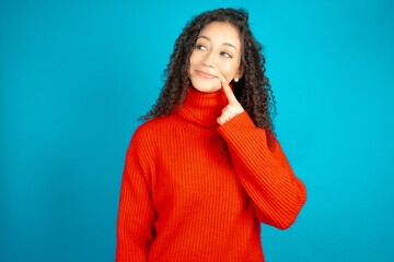 Fototapeta premium Lovely dreamy beautiful teen girl wearing red knitted sweater keeps finger near lips looks aside copy space.