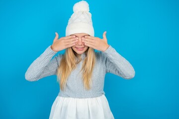 Teen caucasian girl wearing blue knitted sweater and woolly hat covering eyes with hands smiling...