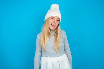 Teen caucasian girl wearing blue knitted sweater and woolly hat winking looking at the camera with...