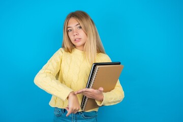 Beautiful kid girl wearing yellow sweater holding notebook In hurry pointing to watch time, impatience, upset and angry for deadline delay.