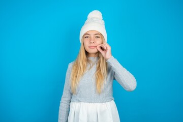 Teen caucasian girl wearing blue knitted sweater and woolly hat mouth and lips shut as zip with...