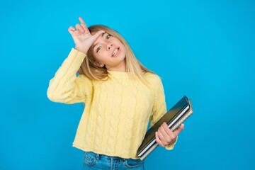 Beautiful kid girl wearing yellow sweater holding notebook making fun of people with fingers on forehead doing loser gesture mocking and insulting.